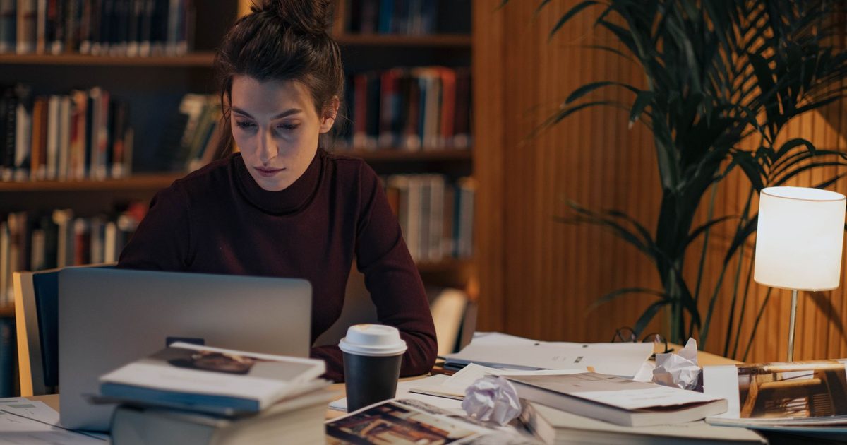 Женщина работает за столом, заваленным раскрытыми книгами, рядом ноутбук